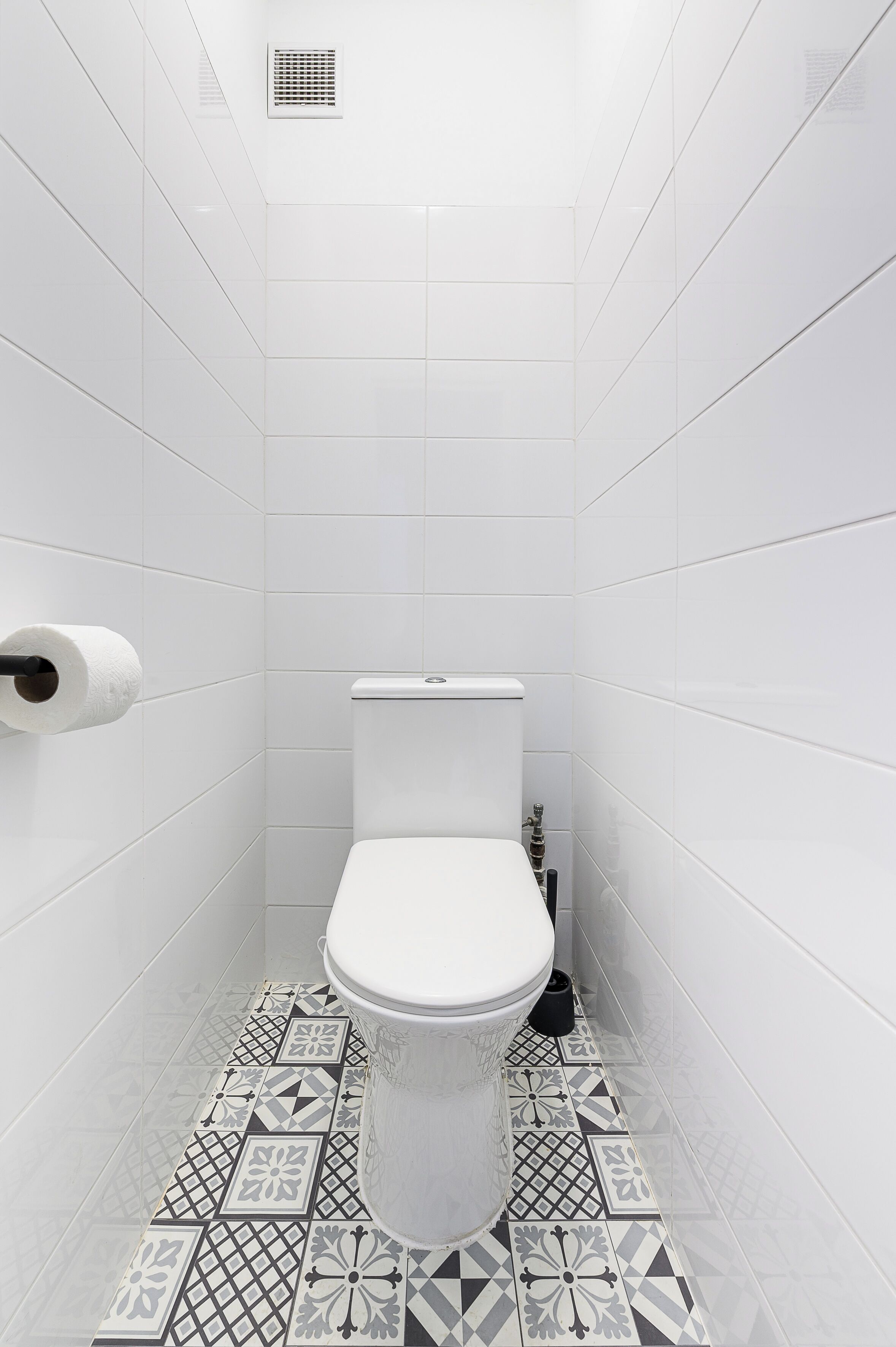Une petite salle de bain aux murs recouverts de carreaux blancs, équipée de toilettes, d’un porte-rouleau de papier toilette et d’un sol carrelé à motifs.