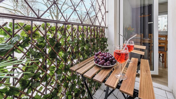 A wooden table with a bowl of grapes and two glasses of red drink, set against a metal lattice with green foliage. - Lumina cozy 3-room apart near the metro (Warsaw)