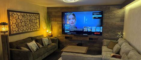 Een woonkamer met een flatscreen televisie aan een houten wand, een donkergroene bank, wanddecoratie met een patroon en een vloerlamp.