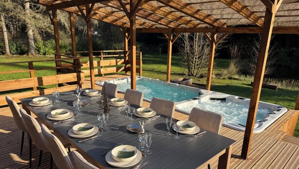 A wooden deck with a table set for six, surrounded by a covered area with a hot tub. - L'écrin de la forêt (Jeanménil)