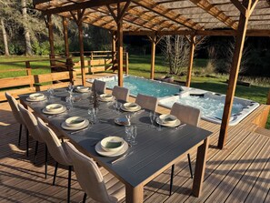 A wooden deck with a table set for six, surrounded by a covered area with a hot tub. - L'écrin de la forêt (Jeanménil)