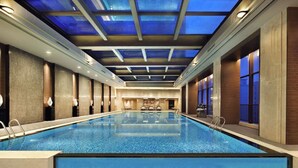 Une grande piscine intérieure dotée d’un toit en verre, entourée de murs en bois et comprenant un espace détente avec des sièges.