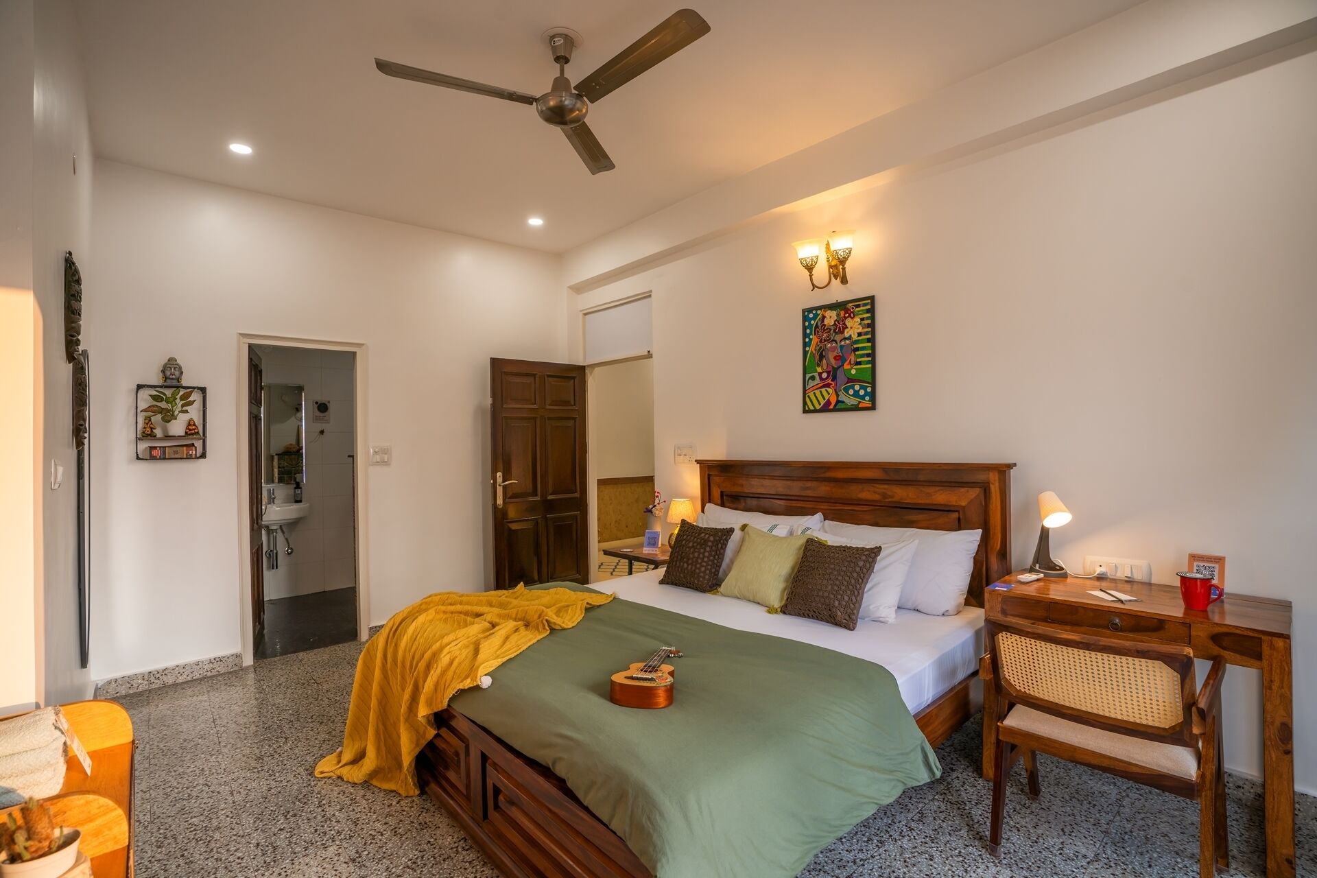 A bedroom with a bed, a desk, a chair, a ceiling fan, a wall-mounted light fixture, and a door leading to a bathroom.