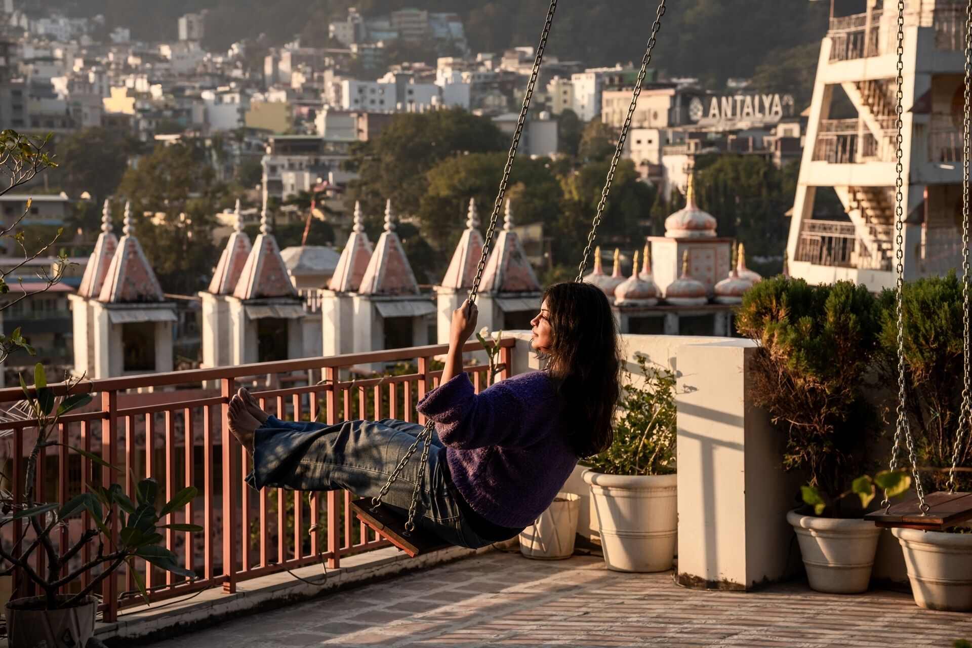 A person on a swing overlooking a cityscape with buildings and a sign reading "ANTALYA".