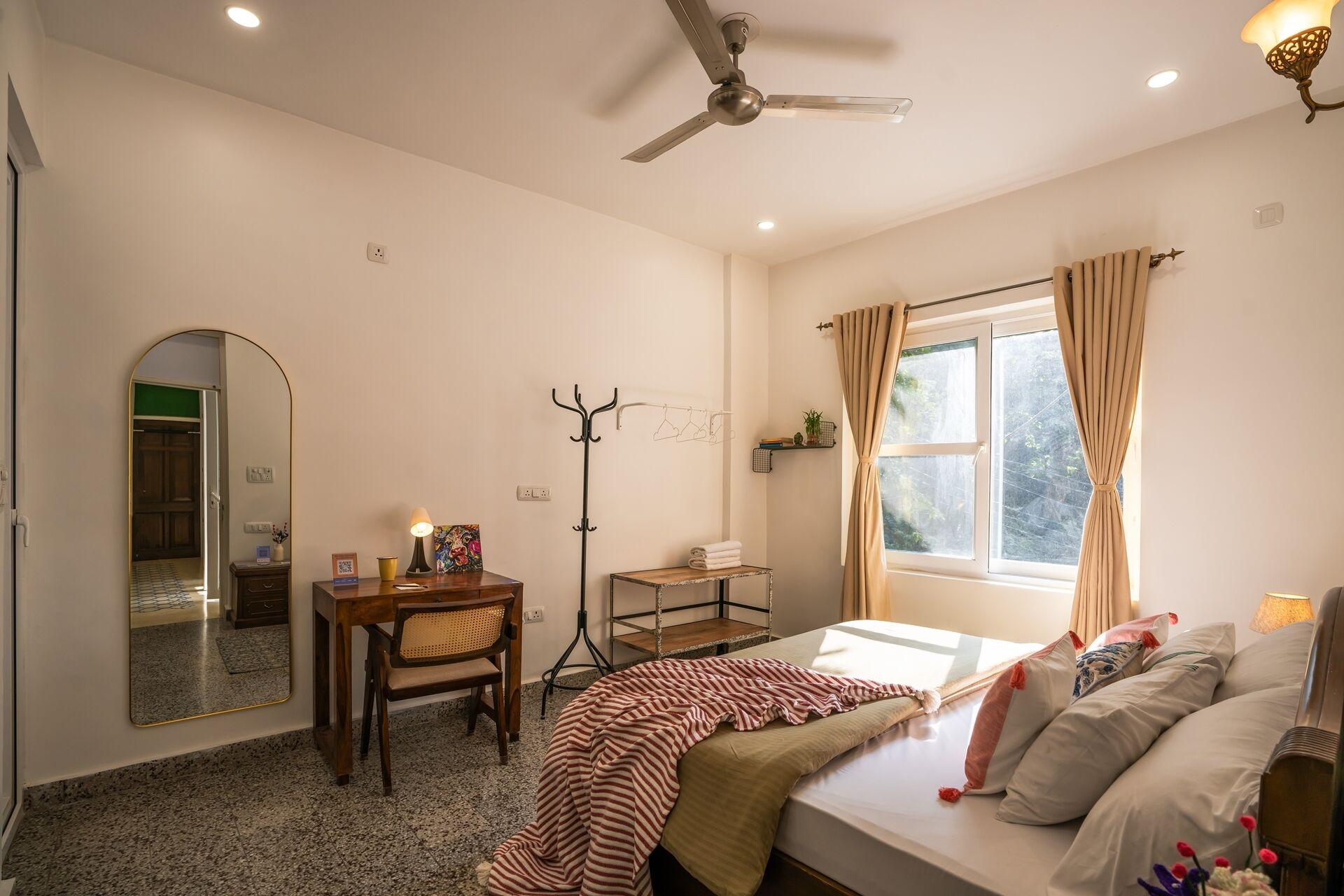 A bedroom with a bed, a desk, a chair, a coat rack, and a window with curtains.