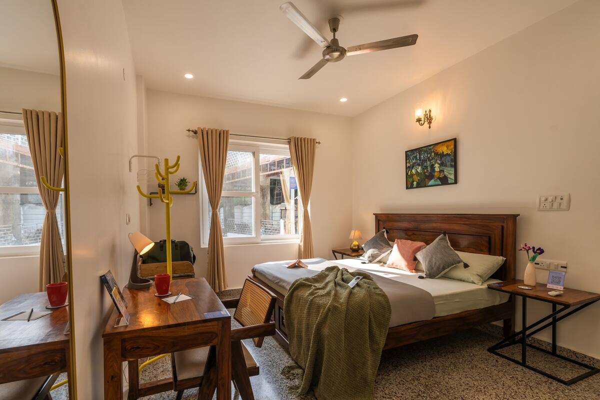 Ein Hotelzimmer mit Bett, Schreibtisch, Stuhl, Kleiderhaken, Deckenventilator und einem Fenster mit Vorhängen.