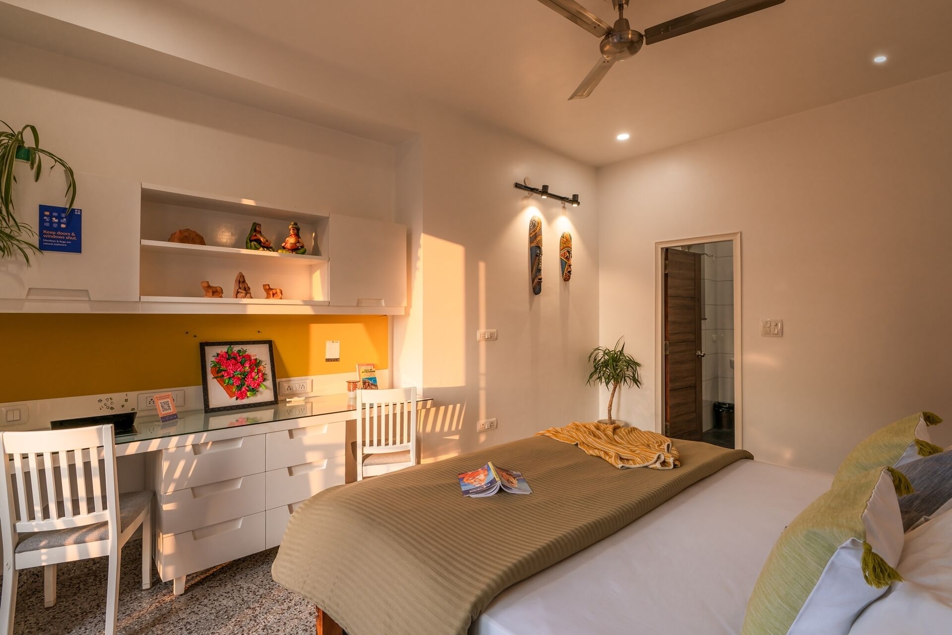 Hotelkamer met een bed, een bureau, een stoel, een klein plankje met beeldjes, een wandlamp en een plafondventilator.