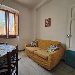A room with a yellow sofa, two patterned pillows, a small table with a floral tablecloth, and a window with curtains. - Grandma Maria's House - The whole house for you (Scilla)