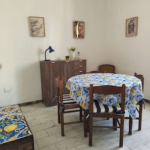 A dining area with a round table covered by a floral tablecloth, surrounded by wooden chairs. There is a wooden cabinet with a blue lamp on top. - Grandma Maria's House - The whole house for you (Scilla)