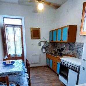A compact kitchen with a dining table, wooden cabinets, a stove, and a window with curtains. - Grandma Maria's House - The whole house for you (Scilla)