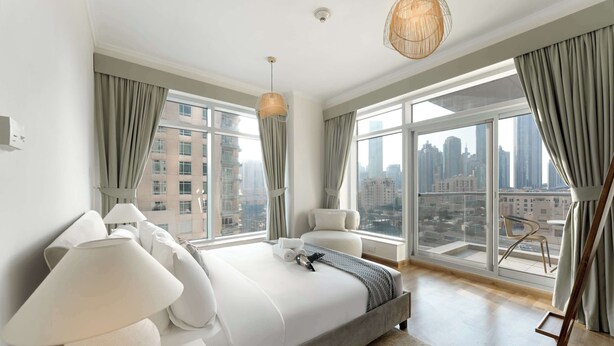 Una camera da letto hotel moderna con un letto grande, numerosi cuscini, un comodino, una lampada e vista sulla città da ampie finestre.
