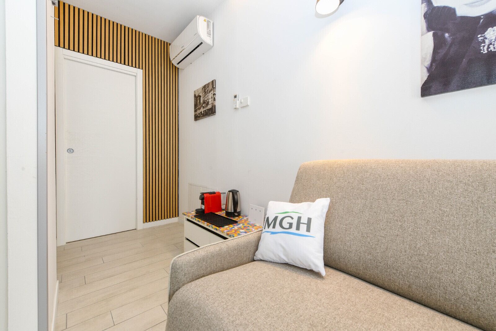 Une petite pièce accueillante, avec un canapé beige, un mur recouvert de lambris en bois, une porte blanche, une petite table sur laquelle se trouvent une bouilloire et une serviette rouge, et un coussin orné des lettres « MGH ».