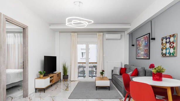 A modern living room with a sofa, a TV, a dining table with red chairs, and a balcony with a view. - Eleanor by halu! - 1-BD Apartment next to Hagia Sophia (Thessaloniki)