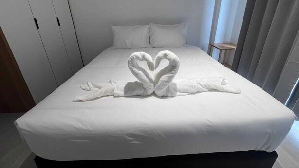 Ein Hotelzimmer mit einem ordentlich bezogenen Bett, einer herzförmigen Handtuchgestaltung und einem Fenster mit Jalousien.