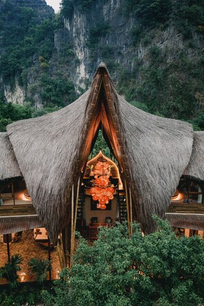 A thatched-roof structure with a central open area, surrounded by lush greenery and a mountainous backdrop. - Lamia Bamboo Retreat Ninh Binh (Tay Hoa Lu)