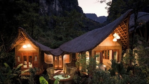 A thatched-roof bungalow with a central courtyard, surrounded by lush vegetation and a mountainous backdrop.