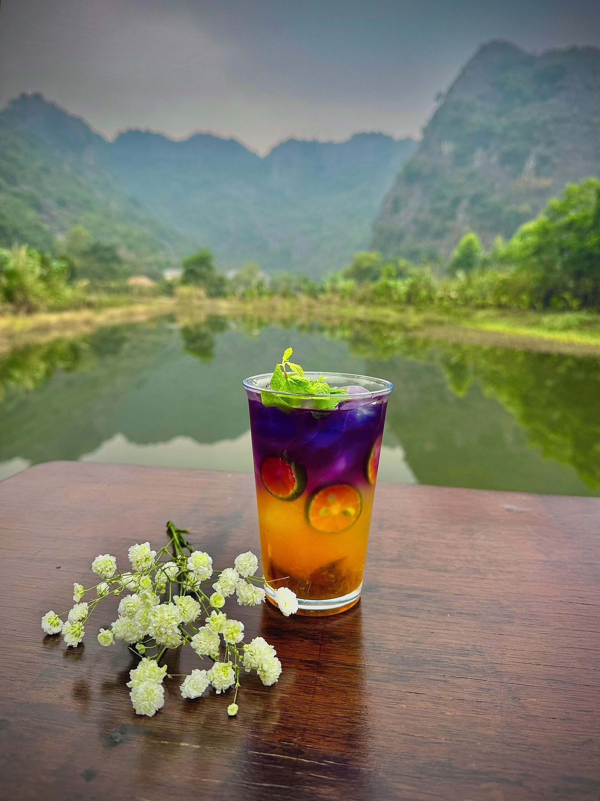 A glass of iced tea with fruit slices and mint on a wooden table with a view of mountains and a lake.