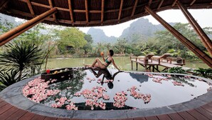 A woman relaxing by a circular pool with lotus flowers, surrounded by lush greenery and a bamboo structure.
