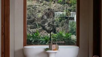 Um banheiro com uma banheira grande, uma pia pequena e uma janela com vista para um jardim rochoso.