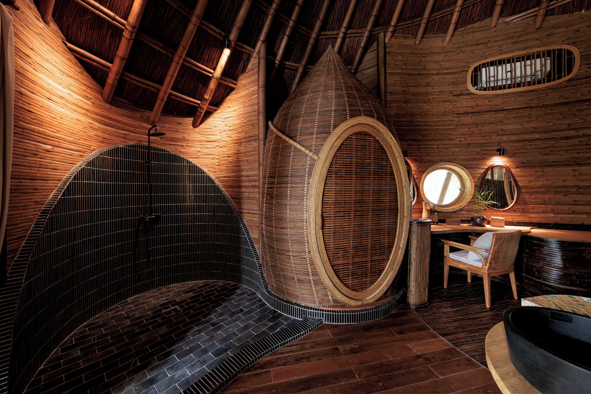 Ein Zimmer mit Holzboden, einem Rundfenster, einem runden Spiegel, einer schwarzen Badewanne und einem Korbstuhl.