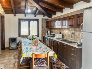 A kitchen with wooden cabinets, a dining table with a floral tablecloth, and a window with curtains. - Casa Gli Allori - Iglesias (Iglesias)