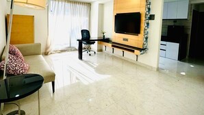 A modern living room with a flat-screen TV mounted on the wall, a sofa, a coffee table, and a dining area with a TV. - Luxury 1401 Pixiestays With Free Parking (Mumbai)