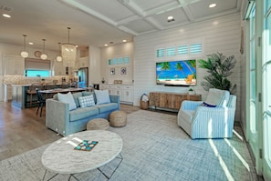 A spacious living room with a large TV, a sofa, a coffee table, and a dining area with a kitchen in the background. - Pool + Boardwalk Access + Steps from the Beach | Villa Blanca by AvantStay (Port Aransas)