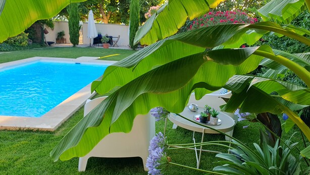 Un espace aménagé au bord de la piscine, avec une petite table et des chaises, entouré d’une végétation luxuriante et donnant sur une piscine à l’eau d’un bleu limpide.