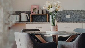 A modern kitchen with a round dining table, two grey upholstered chairs, a vase with white flowers, and a hanging light fixture. - Short walk to the beach and local cafes (Penguin)