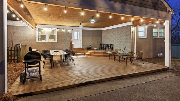 An outdoor patio with a grill, dining table, and chairs, surrounded by a wooden deck and string lights.
