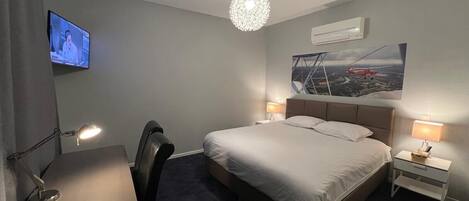 A hotel room with a bed, a desk, a chair, two bedside tables with lamps, a television on the wall, and a painting of a bridge.