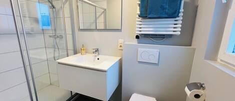 Een moderne badkamer met een douchecabine van glas, een witte wastafel, een toilet en een handdoekrek met handdoeken.