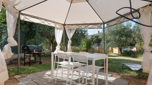Un espacio de patio cubierto con mesa y sillas, una parrilla y vista al jardín.