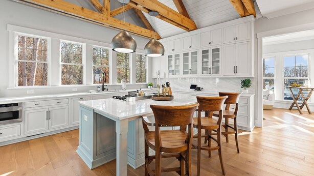 A spacious kitchen with white cabinets, a large island with bar stools, wooden beams on the ceiling, and large windows. - Waterfront Estate + Dock + Pool | Game Room | Sleeps 16 | Southern Belle Estate (Mooresville)