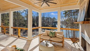 A covered outdoor seating area with a wooden ceiling fan, a sofa with cushions, and a view of a forest and water body. - Waterfront Estate + Dock + Pool | Game Room | Sleeps 16 | Southern Belle Estate (Mooresville)