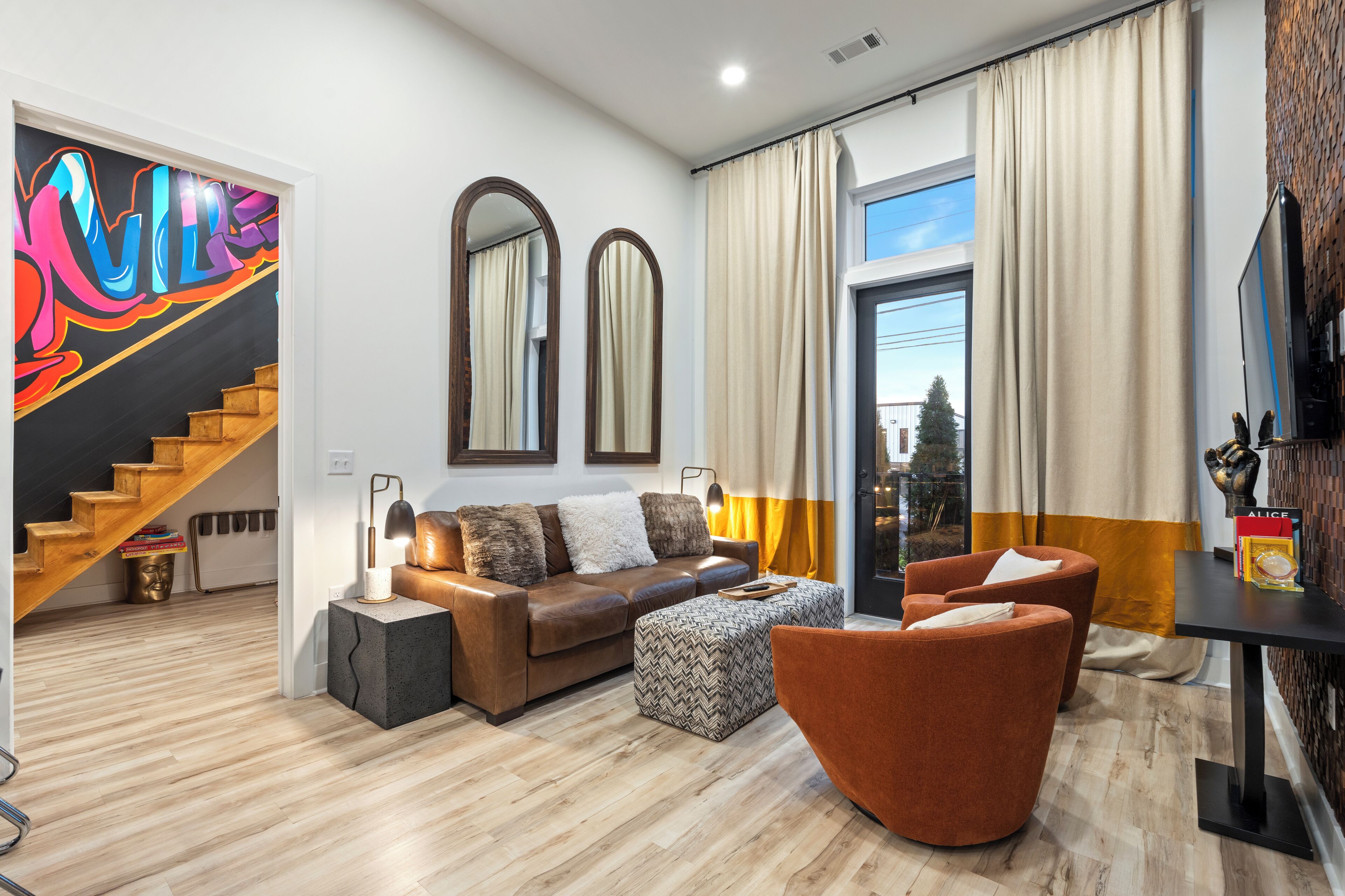 A modern living room with a sofa, ottoman, and armchairs, a staircase with a colorful mural, and a large window with curtains.