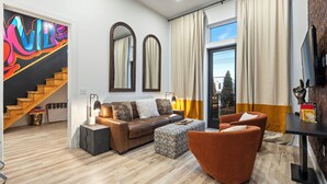 Un salon moderne avec un canapé, un pouf et des fauteuils, un escalier orné d’une fresque colorée et une grande fenêtre avec des rideaux.