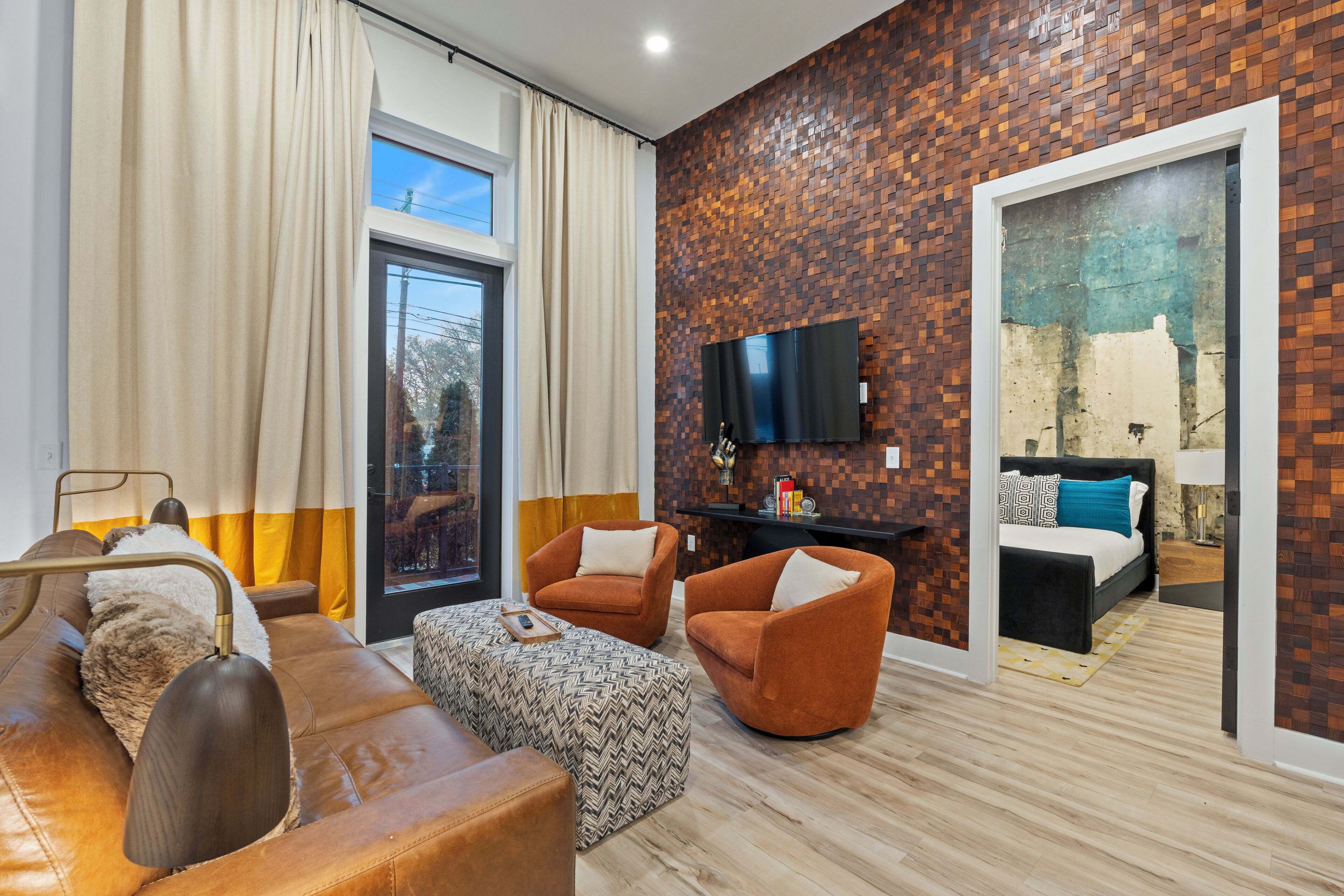 A modern living room with a sofa, armchair, ottoman, and a flat-screen TV mounted on a brick-patterned wall.