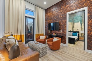 Ein modernes Wohnzimmer mit einer Couch, einem Sessel, einem Hocker und einem Flachbildfernseher, der an einer Wand mit Ziegeloptik angebracht ist.