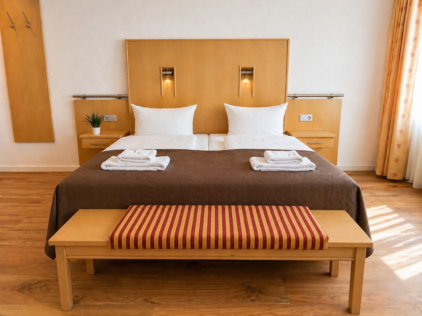 A double bed with a brown bedspread, two white pillows, two bedside tables with lamps, a small plant, and a bench with a striped cushion.