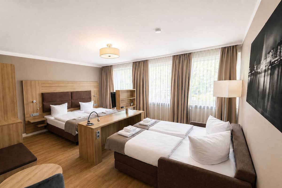 Una camera d'albergo con due letti, una scrivania, una sedia e un'ampia finestra con tende.