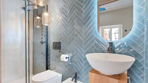 Une salle de bain moderne avec un pare-chocs en carreaux disposés en chevron, des toilettes blanches, un lavabo sur colonne et une douche à parois de verre.