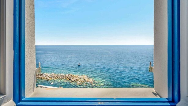 Une vue à travers une fenêtre à cadre bleu, donnant sur une mer calme, une côte rocheuse et un ciel dégagé.