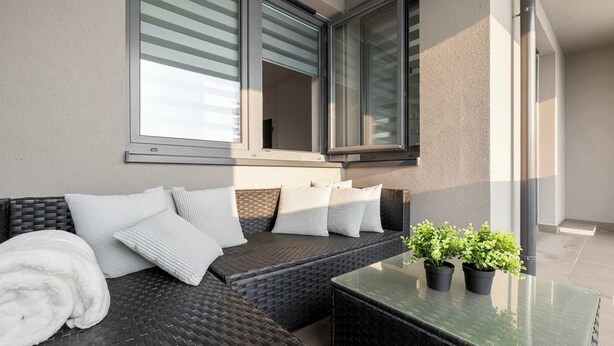 Ein Balkon mit einer Rattan-Couch, weißen Kissen und zwei Blumentöpfen.