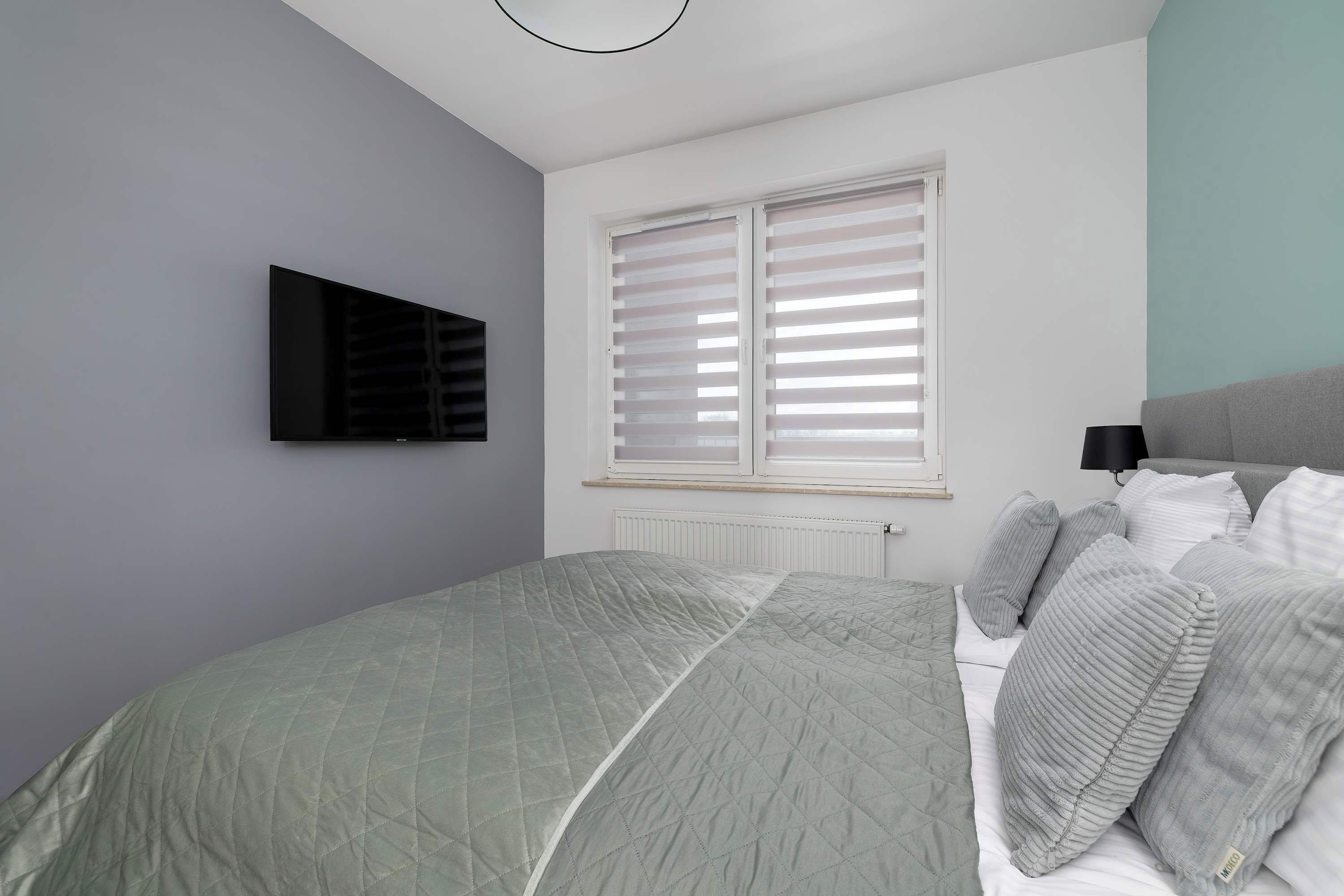 Ein Schlafzimmer mit einem Bett, einem an der Wand befestigten Fernseher und einem Fenster mit Jalousien.