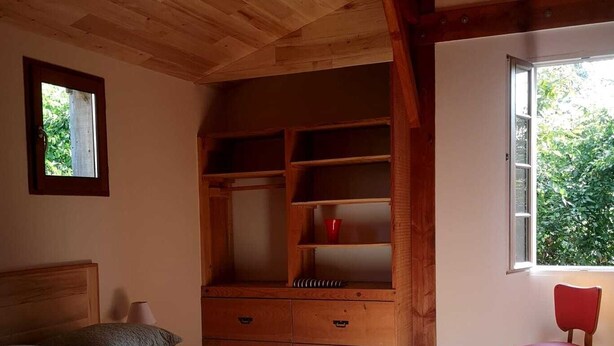 A bedroom with a wooden bed, a built-in wardrobe, a window with a view of greenery, and a red chair.