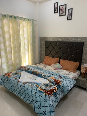 A bedroom with a bed, curtains, a headboard, pillows, a bedside table, and wall art. - Hotel Maitri (Dhanaulti)