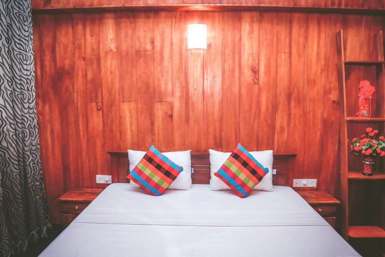 A wooden-paneled room with a bed, two colorful pillows, a nightstand, a shelf with flowers, and a patterned curtain.