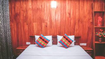 A wooden-paneled room with a bed, two colorful pillows, a nightstand, a shelf with flowers, and a patterned curtain.