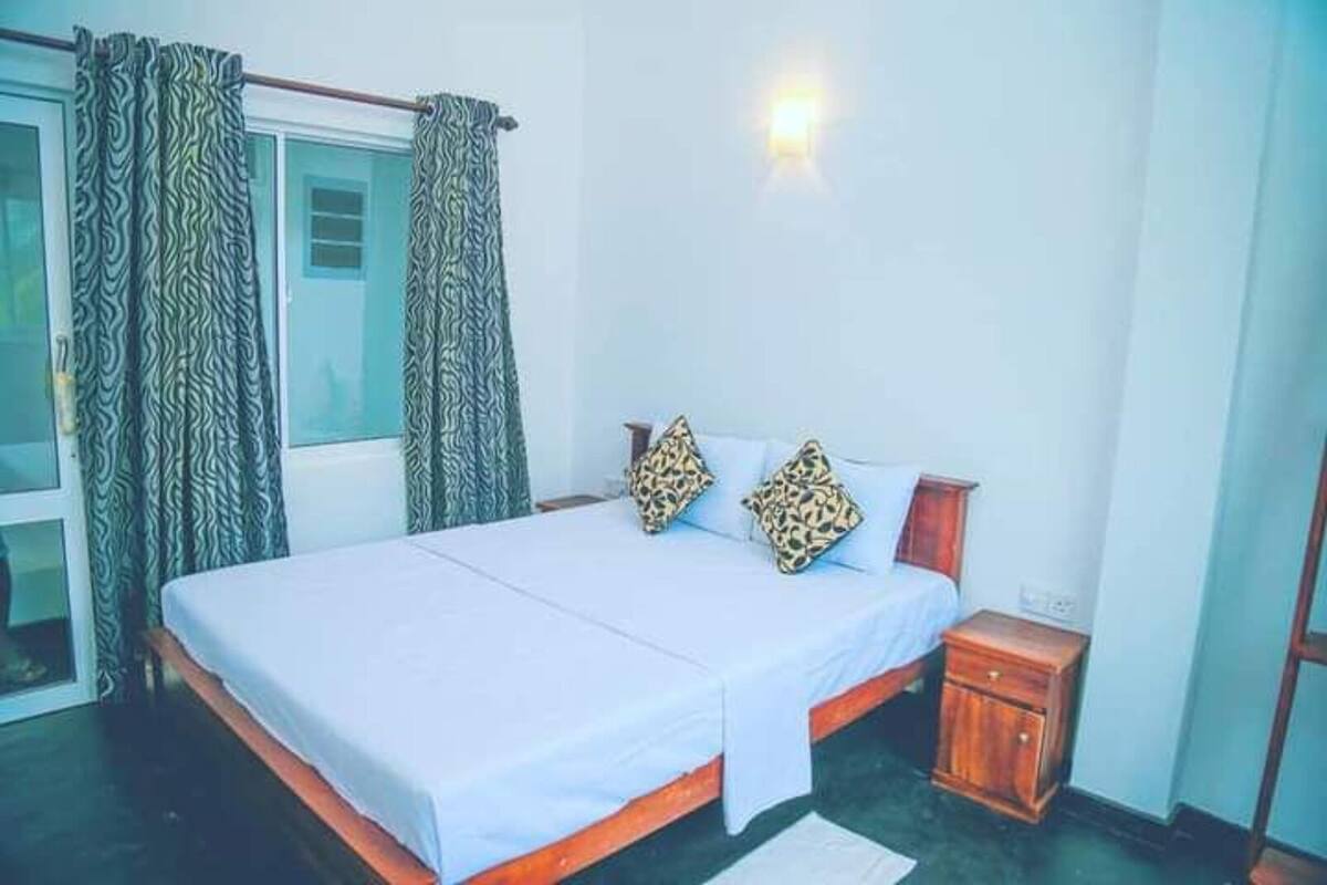 Quarto com cama, cortinas, mesa de cabeceira e luz acoplada à parede.